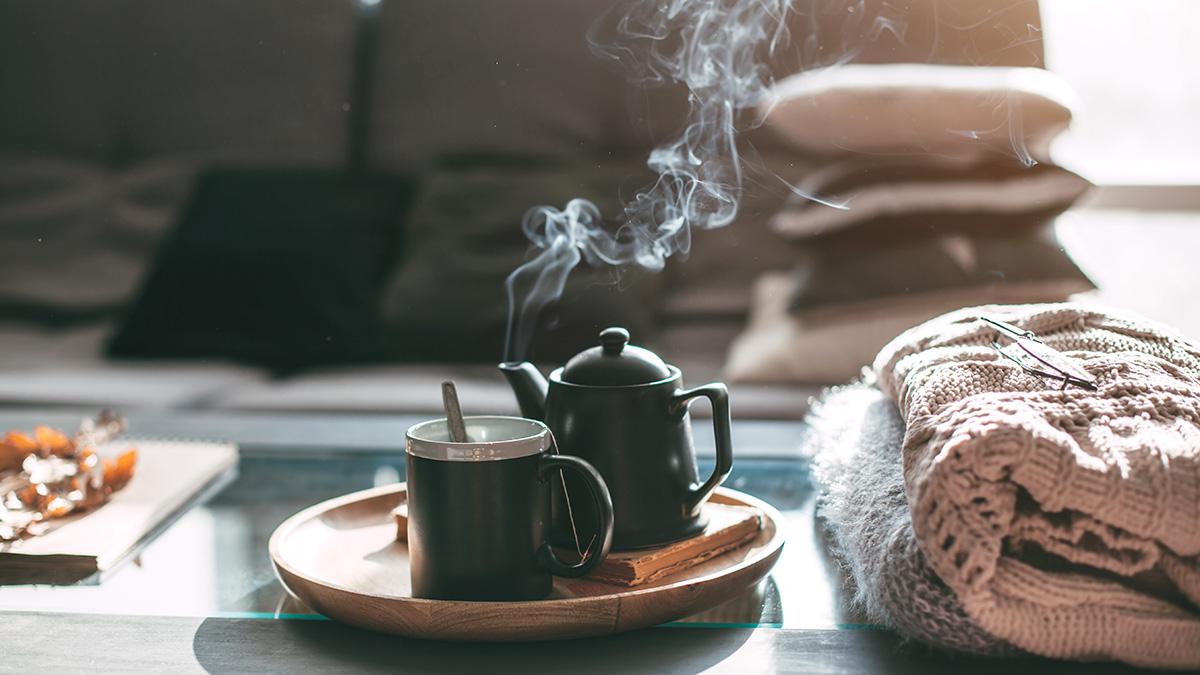 Article Cards Featured Image Still life details in home interior of living room. Sweaters and cup of tea with steam on a serving tray on a coffee table. Breakfast over sofa in morning sunlight. Cozy autumn or winter concept.